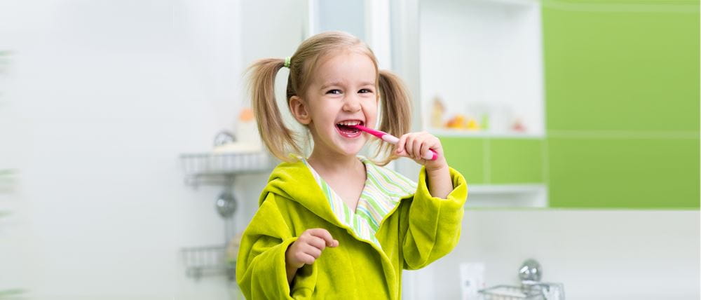 person indoor woman girl brushing young holding teeth looking standing hair front mirror kitchen toothbrush green using shirt wearing table phone sink brush room doing