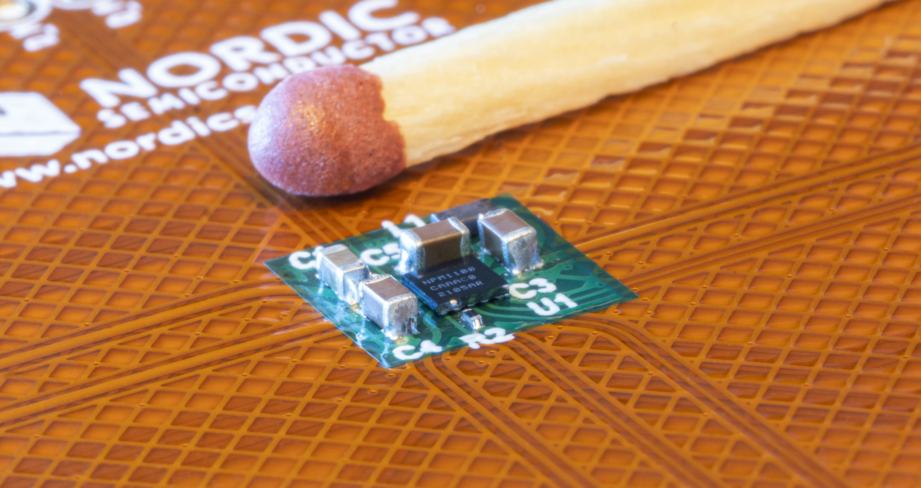 A picture of a very small IC surronded by small capacitors and an inductor, with a matchstick head for scale